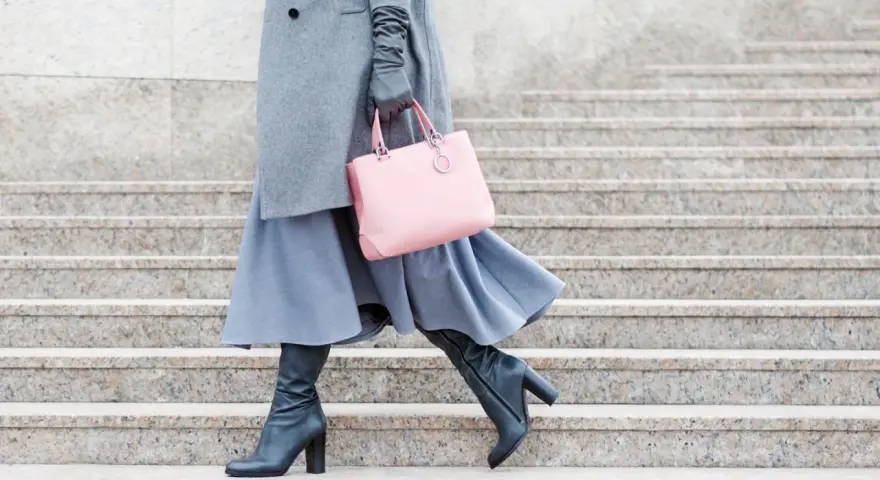 Stilvoll **Kleider im Winter tragen**: Grauer Mantel, blauer Rock, schwarze Stiefel und eine rosafarbene Tasche auf Steinstufen.