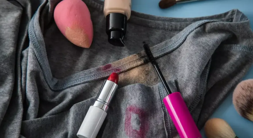 Lippenstiftflecken entfernen: Roter Lippenstift, Mascara, Foundation und Make-up-Schwamm auf grauer Kleidung.