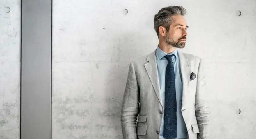 Stilvolles herren outfit sportlich elegant ab 50: Mann mit grauem Sakko, blauem Hemd und Krawatte vor Betonwand.