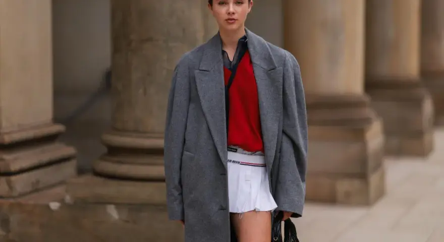 Ein junges Model präsentiert einen preppy look mit grauem Mantel, rotem Pullover und weißem Faltenrock.
