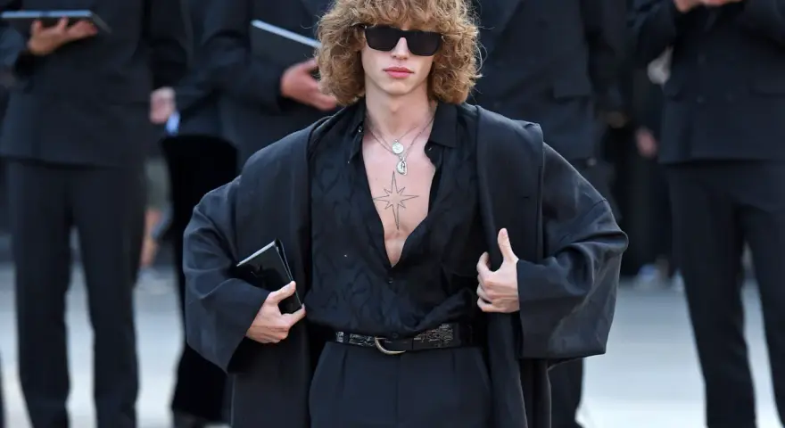Ein Mann mit lockigem Haar und Sonnenbrille trägt eine schwarze, offen geknöpfte Bluse, weite Hosen und einen Mantel. Die Kleidung wirkt typisch 80er Jahre.