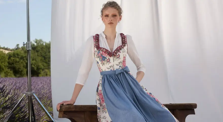 Ein Dirndl im floralen Muster mit blauem Rock, ein moderner Dirndl-Trend, posiert vor Lavendelfeld-Kulisse.