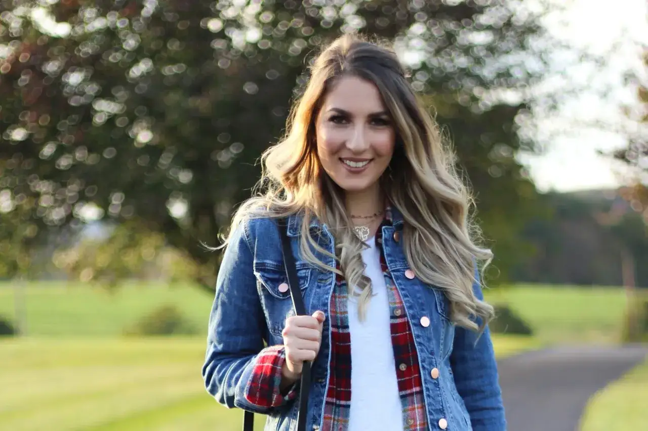 Frau mit blonden Haaren trägt eine Jeansjacke, die sie lässig mit einem karierten Hemd und einem weißen T-Shirt kombiniert.