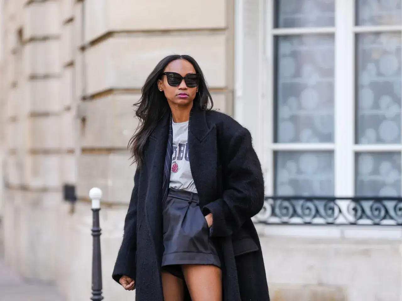Frau in schwarzem Mantel und l&auml;ssigem T-Shirt, die zeigt, wie man ledershorts kombinieren kann. Sonnenbrille und dezentes Make-up runden den Look ab.