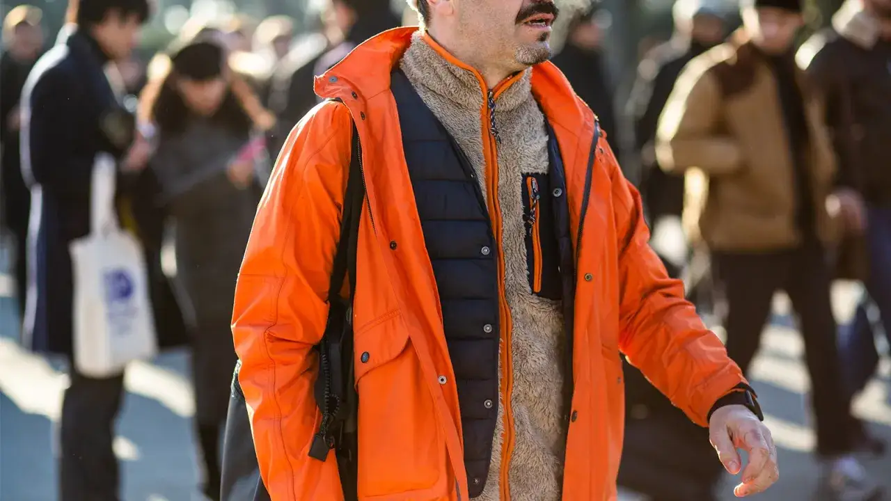 Mann im gorpcore style mit leuchtend oranger Jacke, schwarzer Weste und flauschigem Fleece.