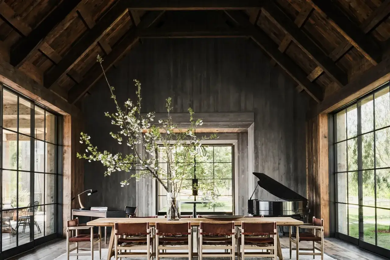 Rustikales Esszimmer mit Holzbalken, Holzwänden, einem langen Holztisch mit Ledersesseln, einem Flügel und einem blühenden Kirschzweig. Ein Hauch von Cottagecore-Charme.