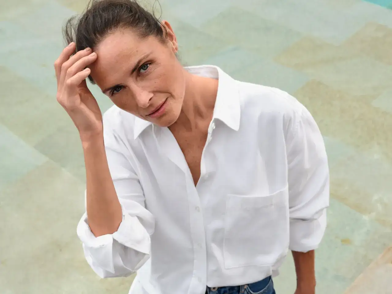 Eine Frau mit 50, die selbstbewusst in die Kamera blickt, ihre Hand leicht am Kopf. Sie trägt ein weißes Hemd und Jeans.