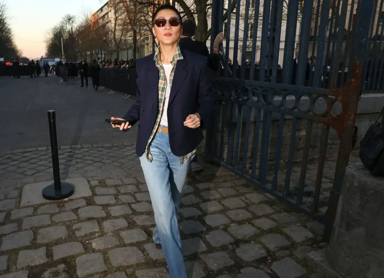 Eine Frau mit Sonnenbrille trägt blaue Jeans, ein kariertes Hemd und einen dunkelblauen Blazer. Ein Beispiel, wie man blaue Jeans kombinieren kann.
