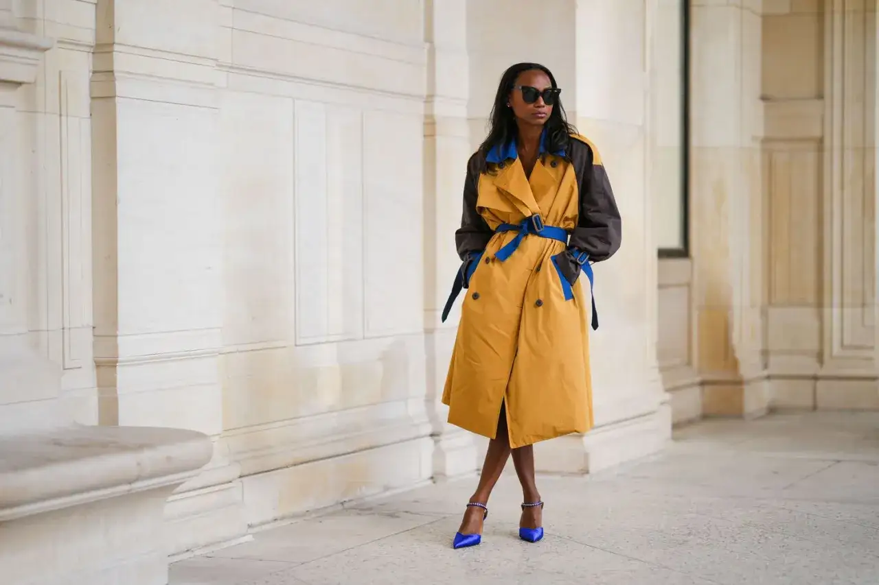Moderne Frau mit Sonnenbrille, die einen gelb-braunen Trenchcoat mit blauem G&uuml;rtel und passenden Pumps kombiniert.