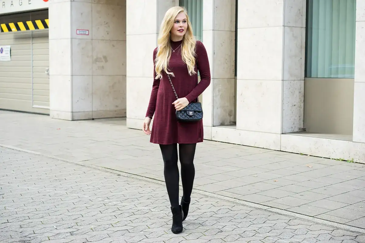 Eine Frau tr&auml;gt ein weinrotes Kleid mit langen &Auml;rmeln und schwarzen Strumpfhosen. Sie beweist, dass man Kleider im Winter tragen kann, und kombiniert sie mit Stiefeletten und einer kleinen Umh&auml;ngetasche.