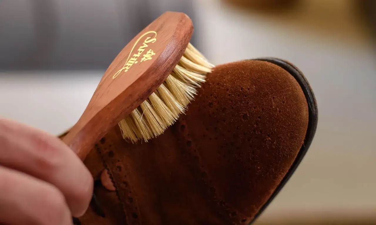 Eine Hand bürstet einen braunen Wildlederschuh mit einer Holz-Bürste. So werden Wildleder Schuhe reinigen und gepflegt.