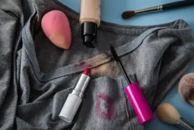 Lippenstiftflecken entfernen: Roter Lippenstift, Mascara, Foundation und Make-up-Schwamm auf grauer Kleidung.