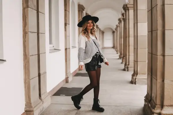 Frau mit Hut und gestreiftem Pullover, die stylische ledershorts kombinieren. Schwarze Stiefel und Strumpfhose runden den Look ab.