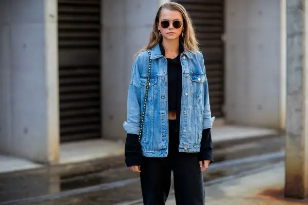 Stylische Frau mit lässiger Jeansjacke kombinieren mit schwarzem Crop-Top und Hose. Sonnenbrille und kleine Tasche runden den Look ab.